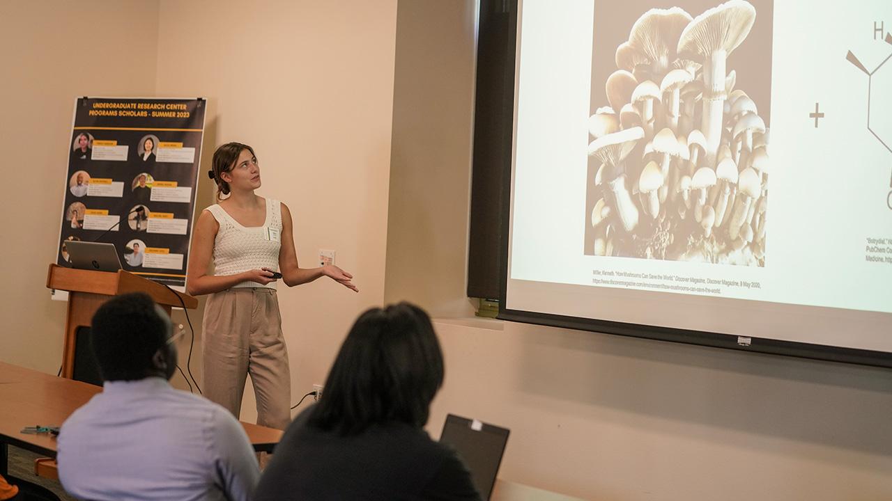 Julia Cook is shown standing in front of a seated audience and gesturing to her research PowerPoint showing an image of mushrooms