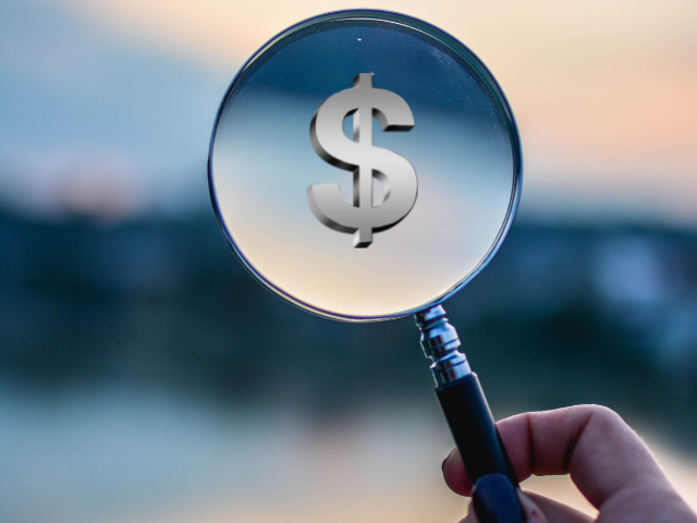 A silver dollar sign appears against an out of focus landscape background through a magnifying glass