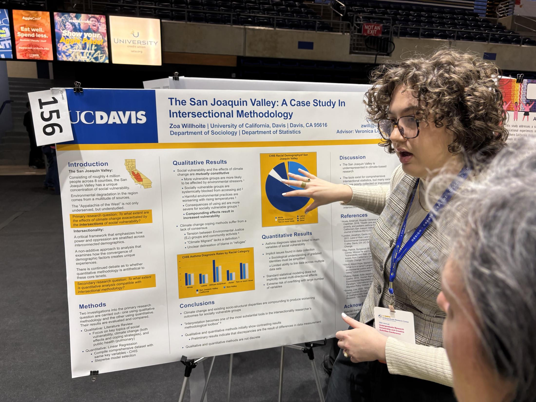 "A student stands next to a research poster titled 'The San Joaquin Valley: A Case Study in Intersectional Methodology' at an academic conference. The student is gesturing toward a pie chart on the poster while explaining the research to an attendee. The poster features sections on introduction, methods, qualitative and quantitative results, and conclusions. It is branded with the UC Davis logo and includes charts and graphs. The student wears glasses, a plaid top, and a conference badge on a blue lanyard.
