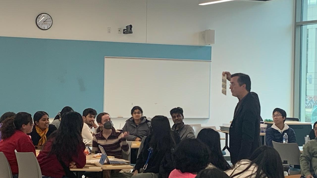 Multiple students sitting in a classroom with some students looking at a faculty member holding up a paper