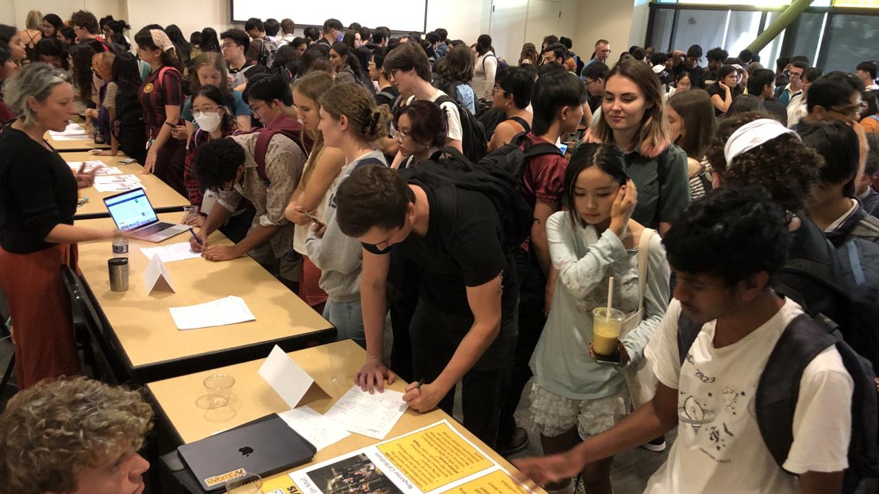 Large group of students gathered around multiple tables set up in rows. 