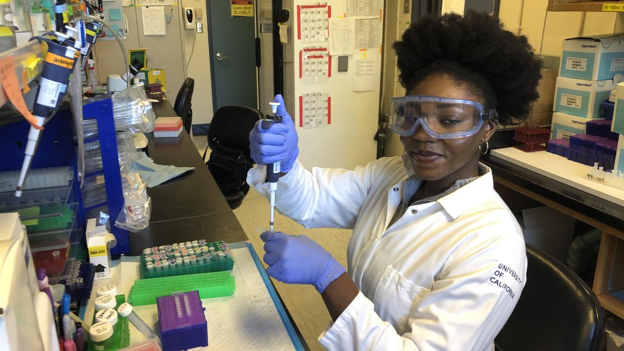 A person wearing a white lab coat, safety goggles, and blue gloves uses a pipette at a lab workstation. The workstation is filled with colorful lab supplies including microcentrifuge tubes, racks, pipette tips, and various containers. The individual is smiling slightly and appears to be in the middle of an experiment.