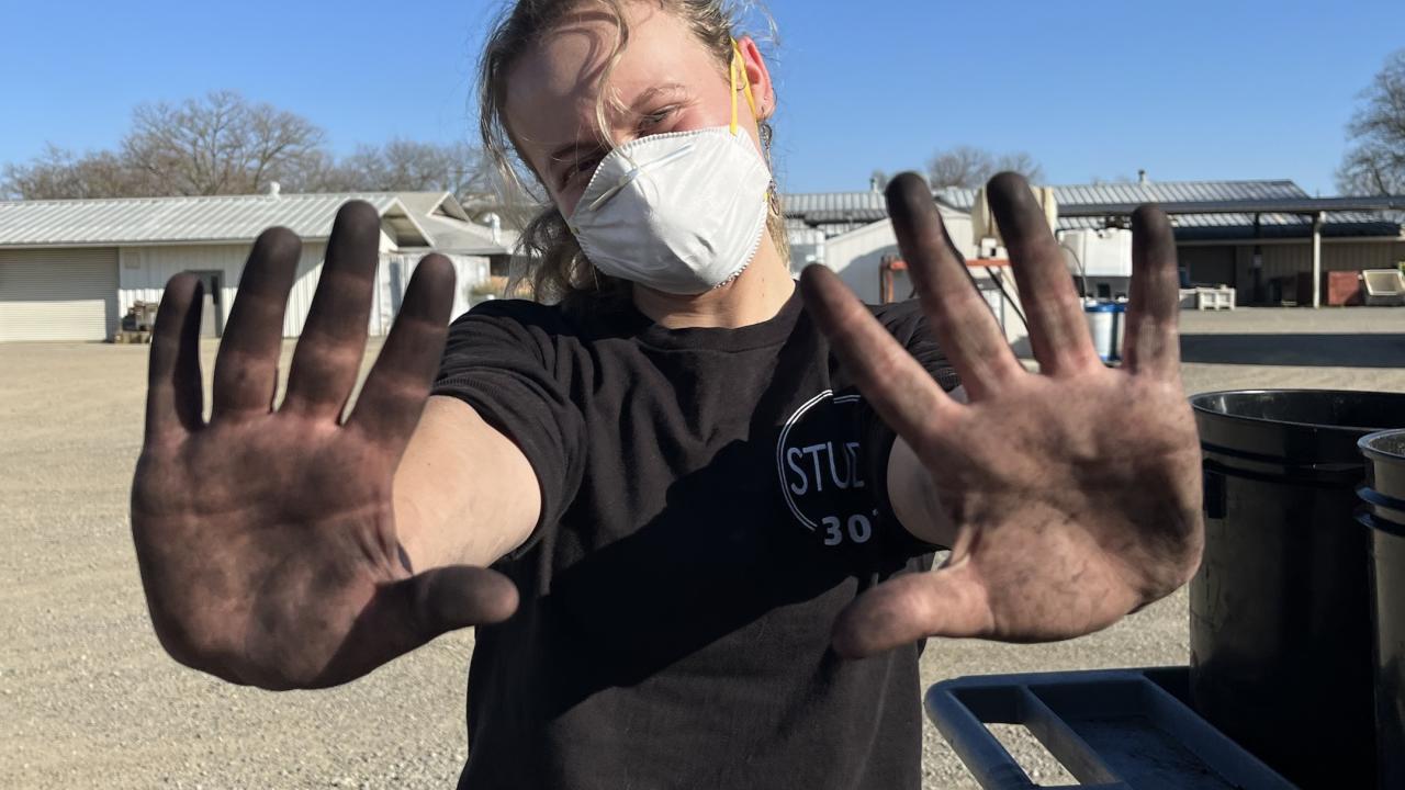 A person wearing a white mask and black T-shirt stands outdoors on a sunny day, holding up both hands covered in black dirt or grease toward the camera. The person has light-colored hair pulled back and is standing in a gravel lot with metal buildings and trees in the background.
