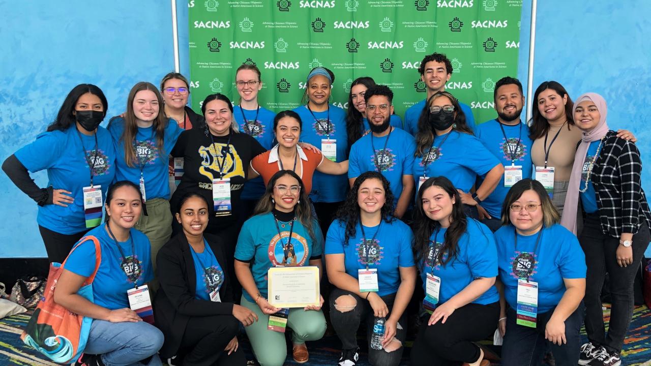 A photo of UC Davis SACNAS conference in Puerto Rico participants, mostly in blue t-shirts with a #thinkbigdiversity design on them, posing in front of a photo backdrop with SACNAS logos