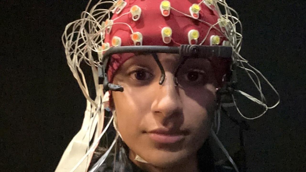 A person wearing a white EEG cap covered with numerous electrodes and wires sits facing the camera. Additional sensors are attached to their face, including one on the forehead with a circular black-and-white target design and another near the cheek. The person is wearing a light-colored sweatshirt and is seated against a dark background,