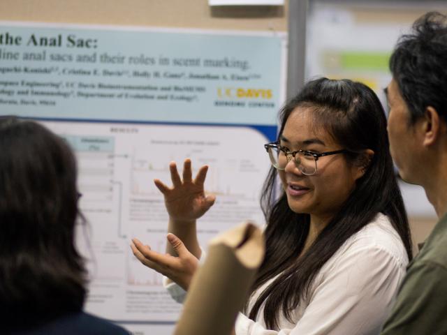 Undergraduate research presenter gesturing in front of research poster at URC Conference