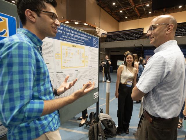 Photo of a conference presenter to the left side of a poster on a poster board presenting to a faculty moderator.  The presenter is gestering and smiling and the moderator is smiling also.  In the background is another smiling presenter on the right side of the poster.