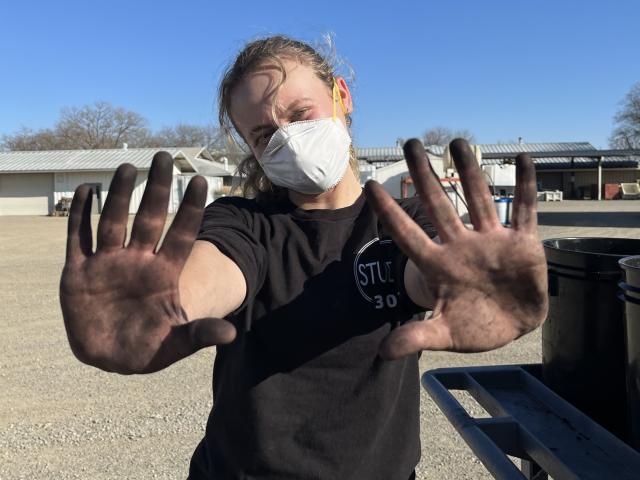 A person wearing a white mask and black T-shirt stands outdoors on a sunny day, holding up both hands covered in black dirt or grease toward the camera. The person has light-colored hair pulled back and is standing in a gravel lot with metal buildings and trees in the background.