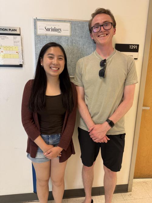 Two smiling individuals stand side by side in a hallway in front of a bulletin board labeled 'Department of Sociology.' The person on the left is wearing a maroon cardigan, dark top, and denim shorts. The person on the right is wearing glasses, a light green T-shirt, black shorts, and a watch, with sunglasses hanging from their shirt. A classroom sign labeled '1291' and an evacuation plan are visible on the wall.