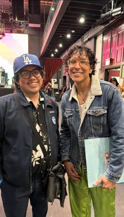 Two people stand smiling indoors at an event venue. The person on the left wears a blue Los Angeles Dodgers cap, glasses, and a dark jacket over a floral shirt. The person on the right wears large orange glasses, a denim jacket with a shearling collar, and shiny green pants, holding a light blue folder. Stage lights, curtains, and event equipment are visible in the background.