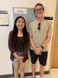Two smiling individuals stand side by side in a hallway in front of a bulletin board labeled 'Department of Sociology.' The person on the left is wearing a maroon cardigan, dark top, and denim shorts. The person on the right is wearing glasses, a light green T-shirt, black shorts, and a watch, with sunglasses hanging from their shirt. A classroom sign labeled '1291' and an evacuation plan are visible on the wall.