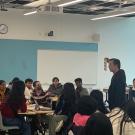 Multiple students sitting in a classroom with some students looking at a faculty member holding up a paper