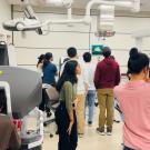 A group of people in a medical or research lab interacting with robotic surgical equipment. One person in the foreground is seated at a Da Vinci surgical console, while another is taking a photo with a smartphone. Several individuals in the background are gathered around a screen displaying a 3D model, while others are observing robotic arms covered in blue surgical drapes. The room is well-lit, with advanced medical technology and monitors mounted on the ceiling.