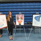 A woman with long brown hair, wearing a black outfit, stands smiling in front of three framed paintings displayed on easels in an indoor exhibition space. The artwork on the left features an abstract composition with splashes of color and floral elements. The middle piece portrays a surreal landscape with layered, colorful hills inside a floating shape against a night sky with trees. The artwork on the right consists of a dark blue abstract form with splattered paint on a white background. The setting inclu