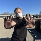A person wearing a white mask and black T-shirt stands outdoors on a sunny day, holding up both hands covered in black dirt or grease toward the camera. The person has light-colored hair pulled back and is standing in a gravel lot with metal buildings and trees in the background.