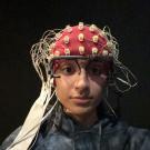 A person wearing a white EEG cap covered with numerous electrodes and wires sits facing the camera. Additional sensors are attached to their face, including one on the forehead with a circular black-and-white target design and another near the cheek. The person is wearing a light-colored sweatshirt and is seated against a dark background,