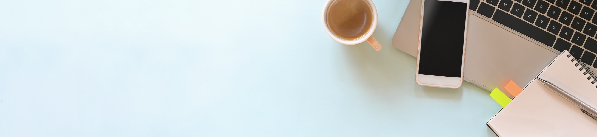 An overhead photo of a corner of a laptop, notebook and a cup of coffee and a smart phone on a light blue surface